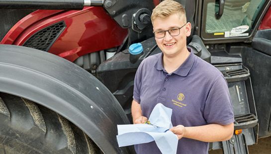 Ein Azubi steht vor einem Traktor und hat Werkzeug in der Hand.